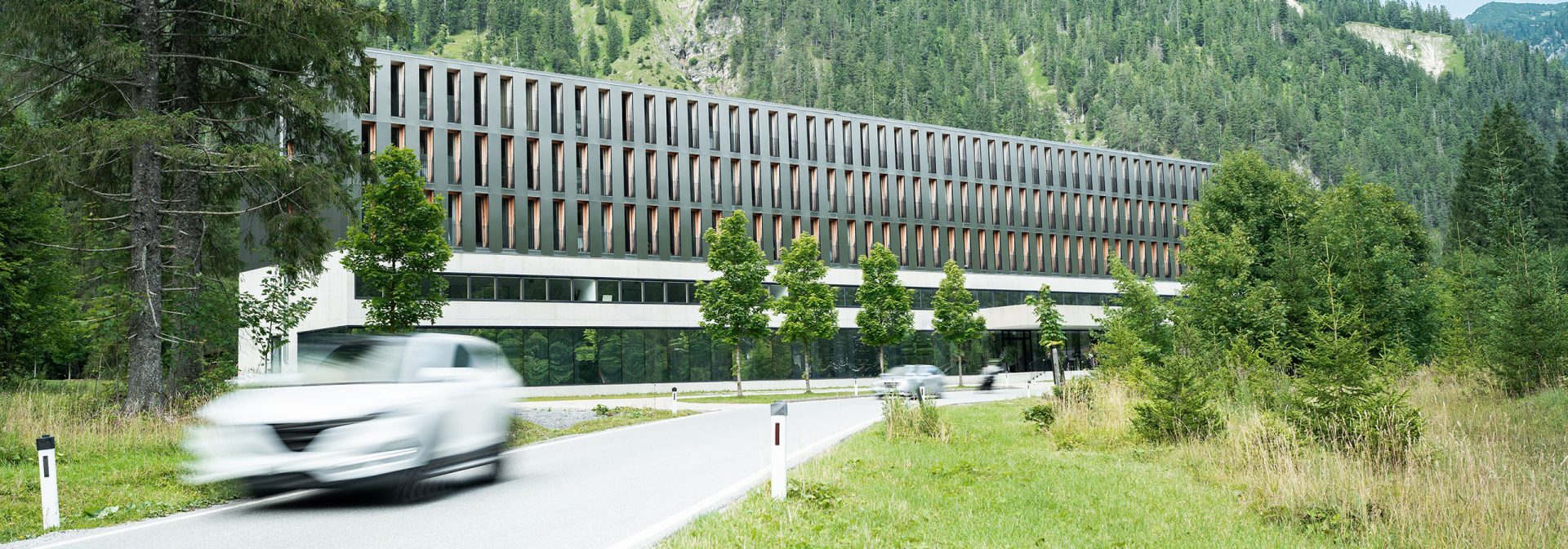 Das Alpenhotel Ammerwald mit einem Berg im Hintergrund und davor eine Straße, auf der sich ein weißes Fahrzeug bewegt.