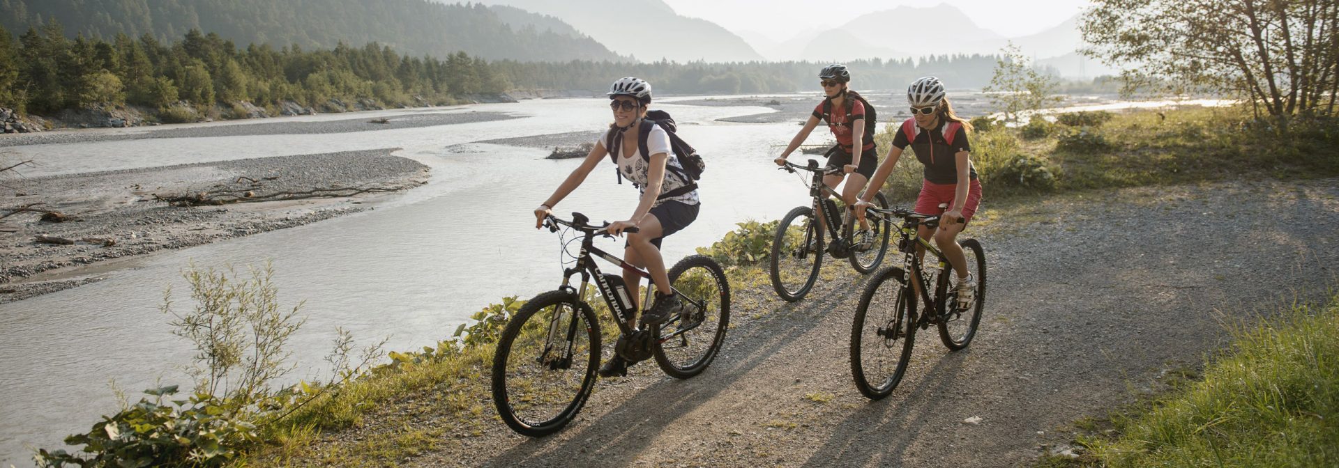 Drei junge Frauen fahren auf Mountainbikes auf einem Schotterweg an einem Fluss entlang. Die Sonne scheint und die drei haben Spaß.