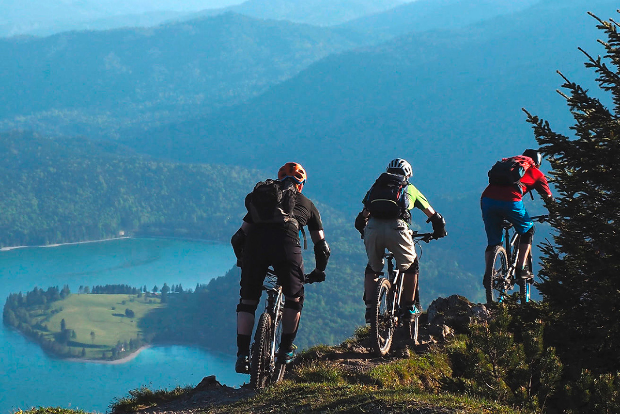 Drei Männer mountainbiken in den Bergen. Im Hintergrund ist ein See.