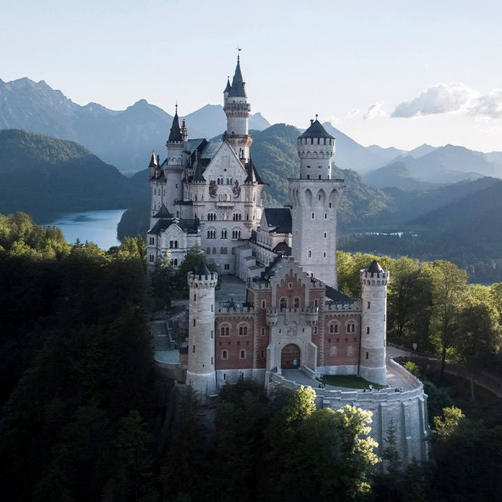 Ein pittoreskes Bild von Schloss Neuschwanstein in den frühen Morgenstunden eines Sommertages. Hinter dem Märchenschloss ist die Bergkulisse der Region. Neben Wäldern und Felsbergen sieht man den Auschnitt eines Sees. 