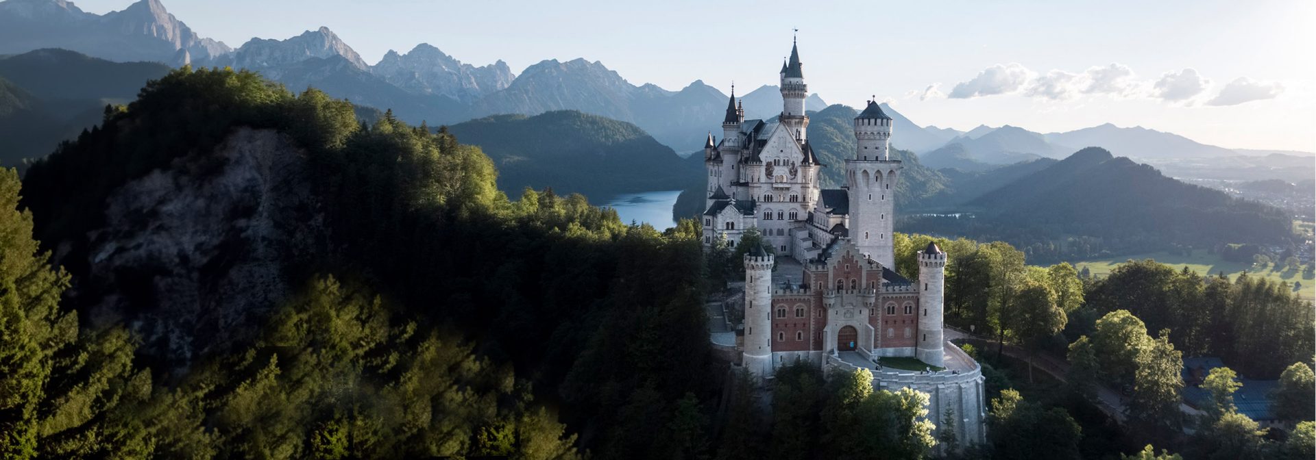 Ein pittoreskes Bild von Schloss Neuschwanstein in den frühen Morgenstunden eines Sommertages. Hinter dem Märchenschloss ist die Bergkulisse der Region. Neben Wäldern und Felsbergen sieht man den Auschnitt eines Sees. 