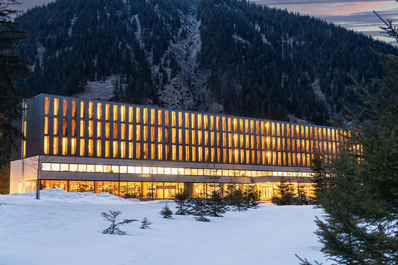 Das Alpenhotel Ammerwald mit einem Berg im Hintergrund und davor eine Straße, auf der sich ein weißes Fahrzeug bewegt.