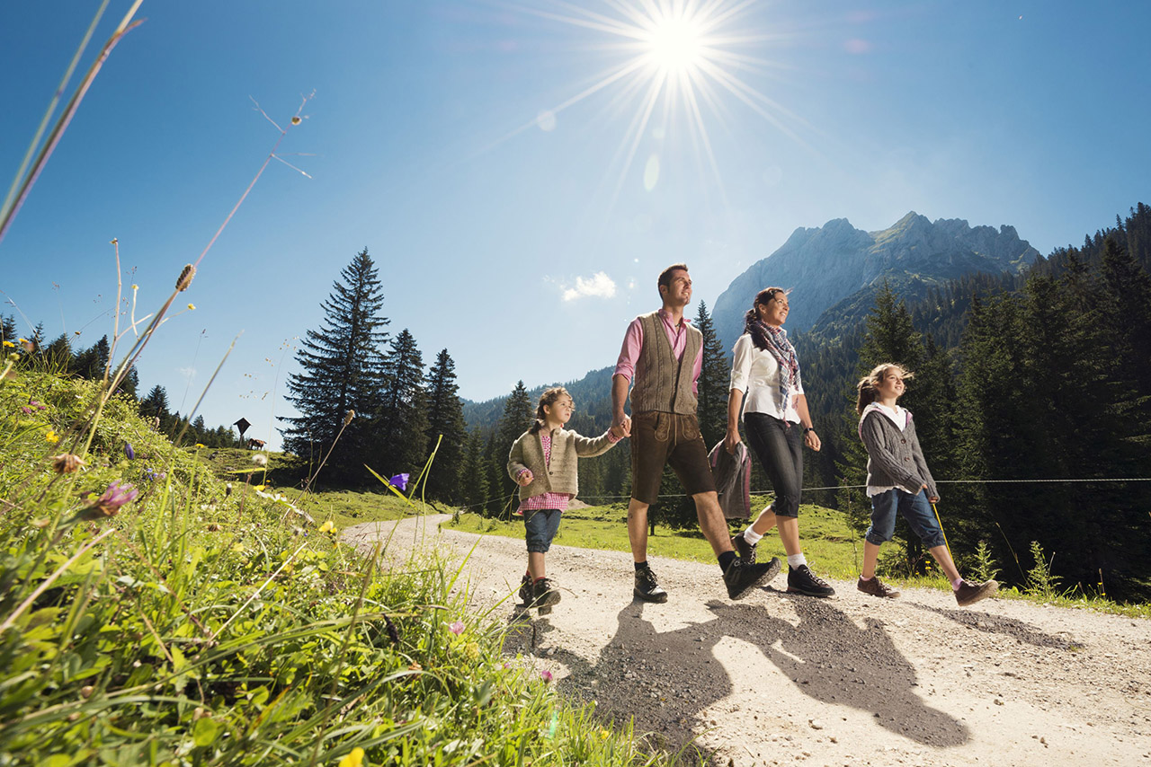 Eine vierköpfige Familie mit Mutter, Vater und zwei Töchtern im Grundschulalter machen eine Sommerwanderung. Die Sonne scheint, auf den Wiesen blühen Sommerblumen und hinter ihnen erstreckt sich das Gebirge.
