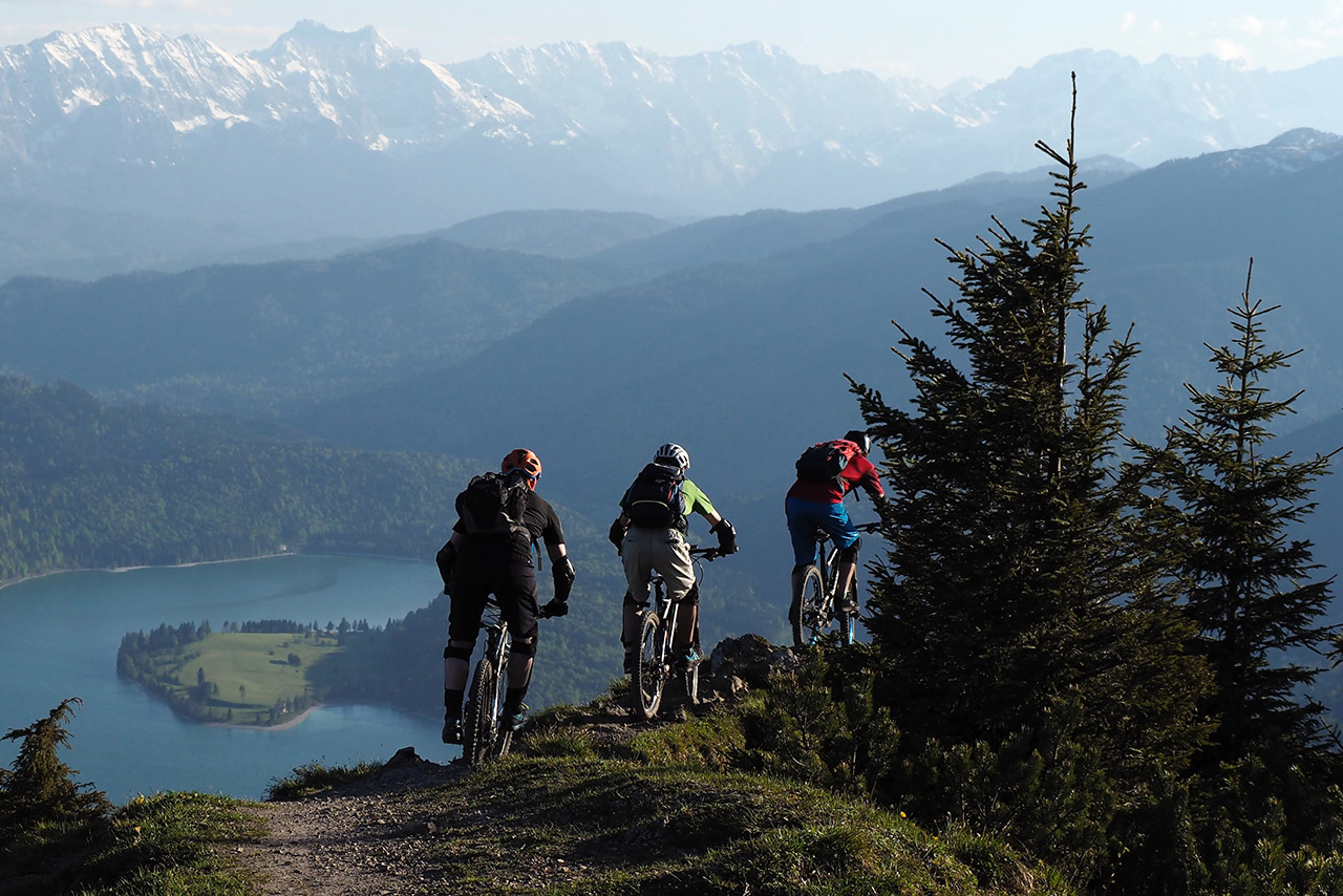 Drei Männer mountainbiken in den Bergen. Im Hintergrund ist ein See.