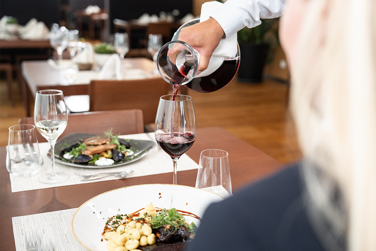 Ein Kellner schenkt einer Frau im Restaurant Rotwein ein. Es steht bereits Essen auf dem Tisch: Ein Teller mit dem Hirschragout, sowie ein Ribeye Steak 350.