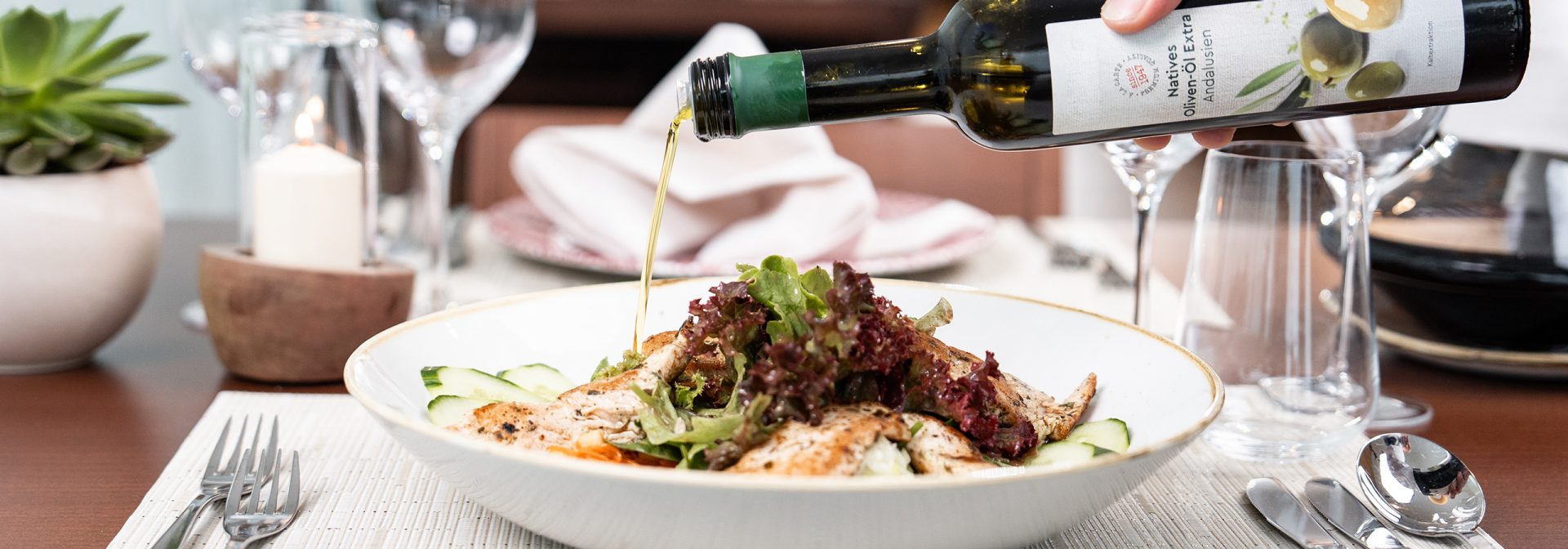 DE: Ein gedeckter Tisch im Restaurant. Jemand gibt Olivenöl über einen Maishähnchenbrustsalat. 