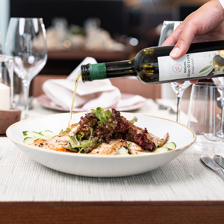 DE: Ein gedeckter Tisch im Restaurant. Jemand gibt Olivenöl über einen Maishähnchenbrustsalat. 