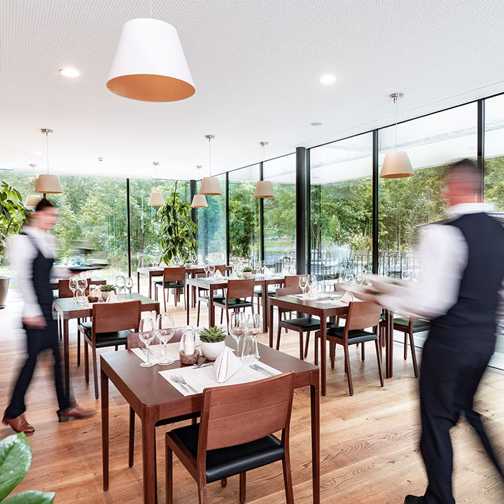 A modern restaurant interior featuring wooden tables and large windows with a view of greenery.
