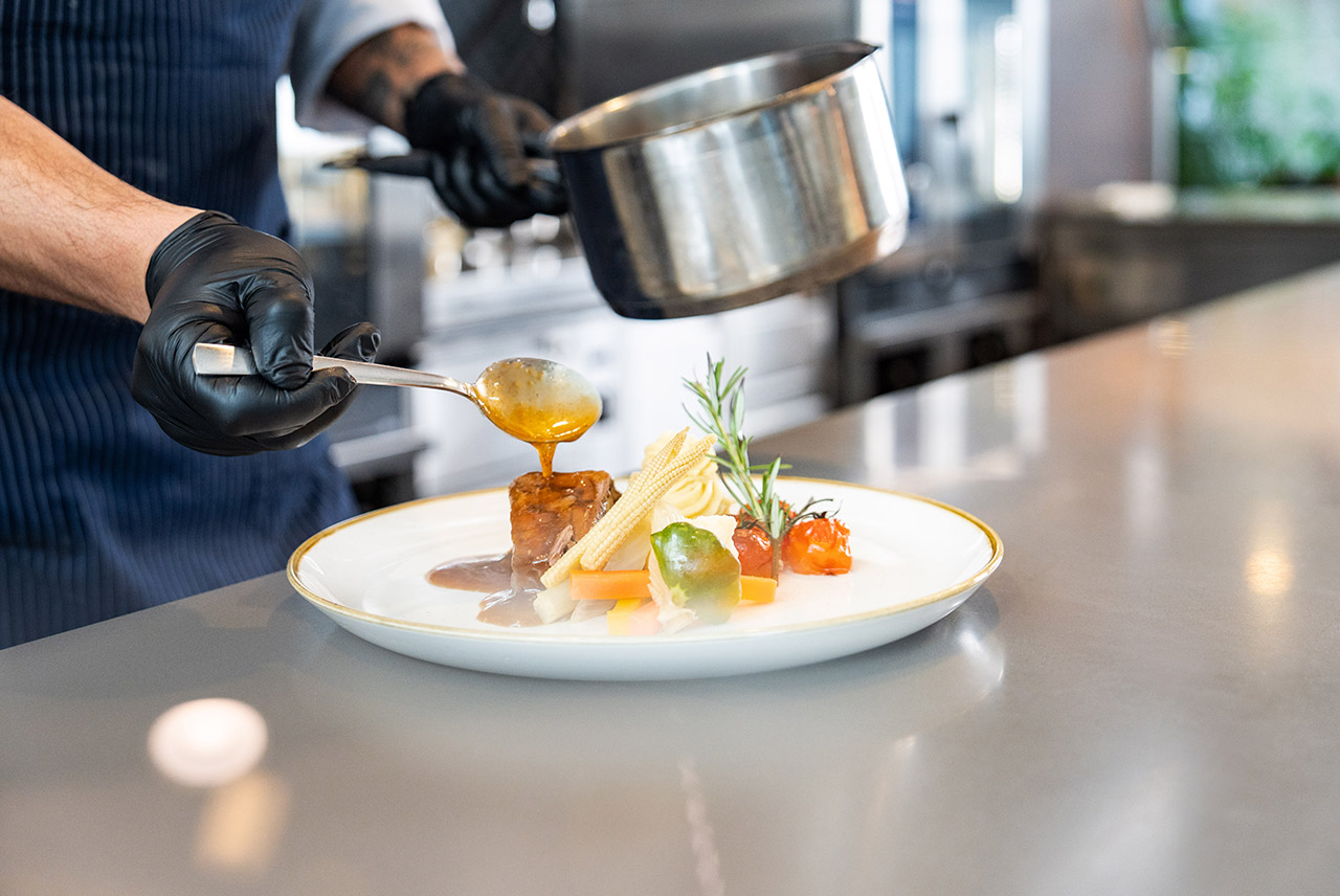 Ein Kellner schenkt einer Frau im Restaurant Rotwein ein. Es steht bereits Essen auf dem Tisch: Ein Teller mit dem Hirschragout, sowie ein Ribeye Steak 350.