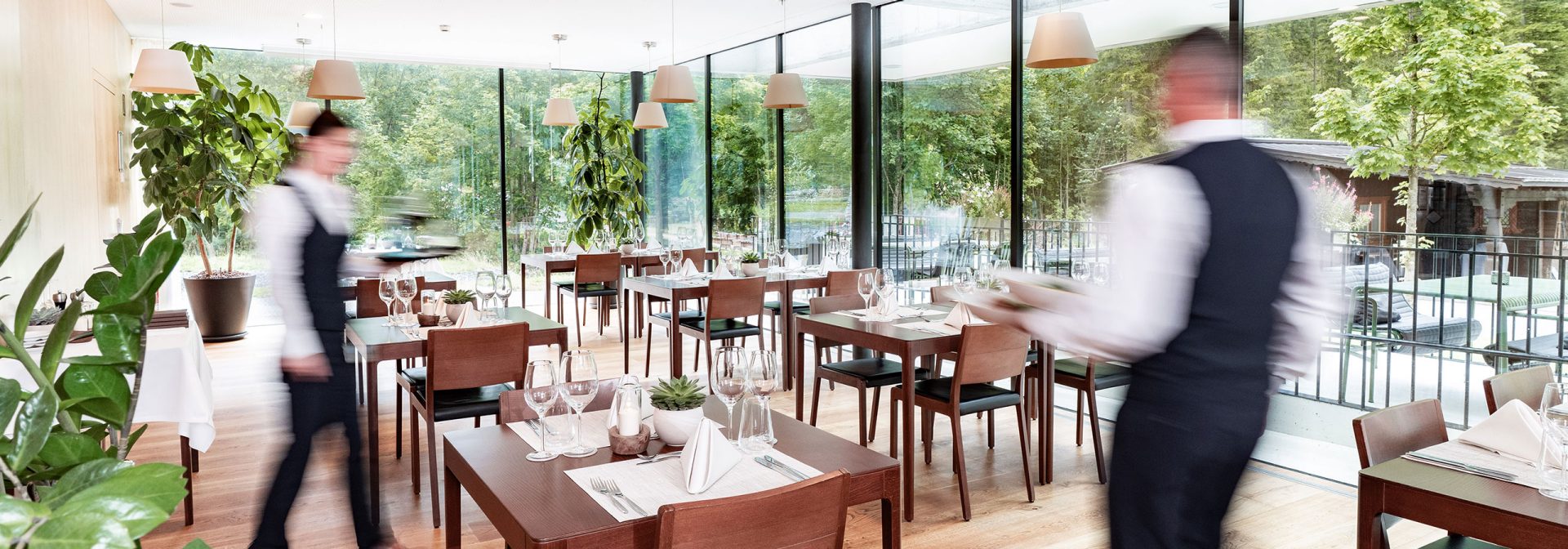 A modern restaurant interior featuring wooden tables and large windows with a view of greenery.