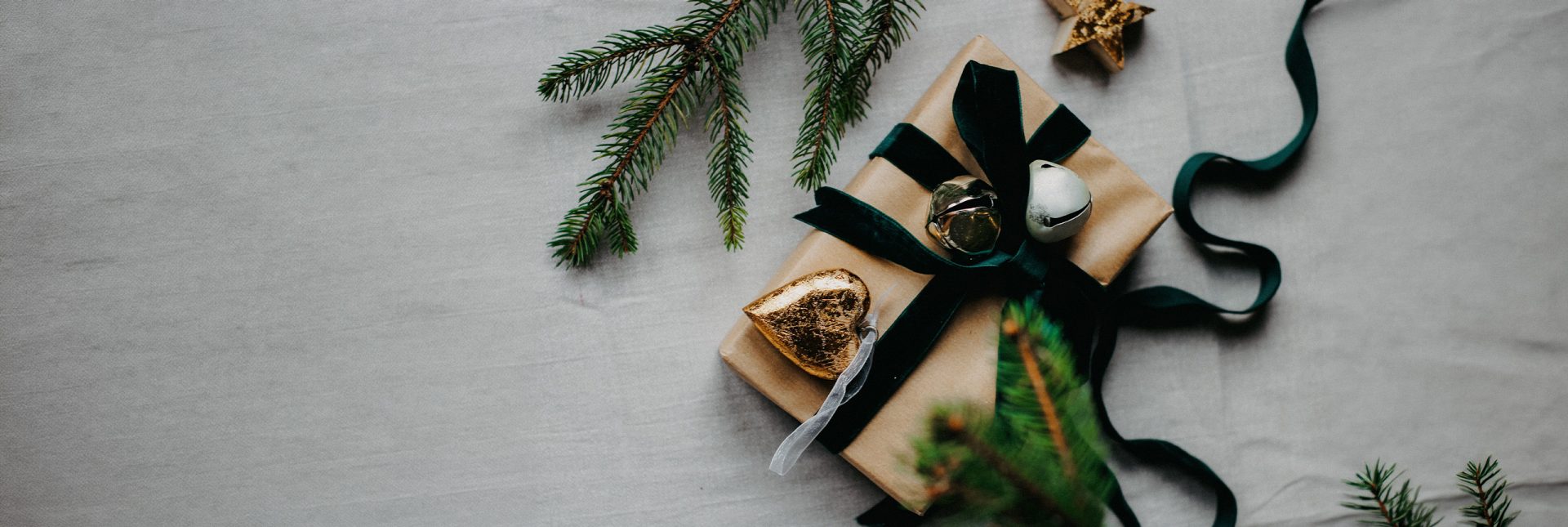 Gift wrapped with black ribbon and gold bell next to fir branch on gray surface.